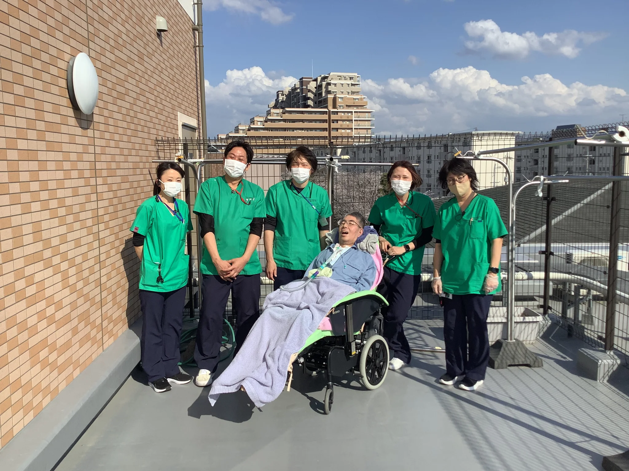 ReHOPE 御殿山南の屋上で、車椅子のご入居者さまを囲んで並ぶ緑色のユニフォームを着たスタッフ5人。青空の下での記念写真
