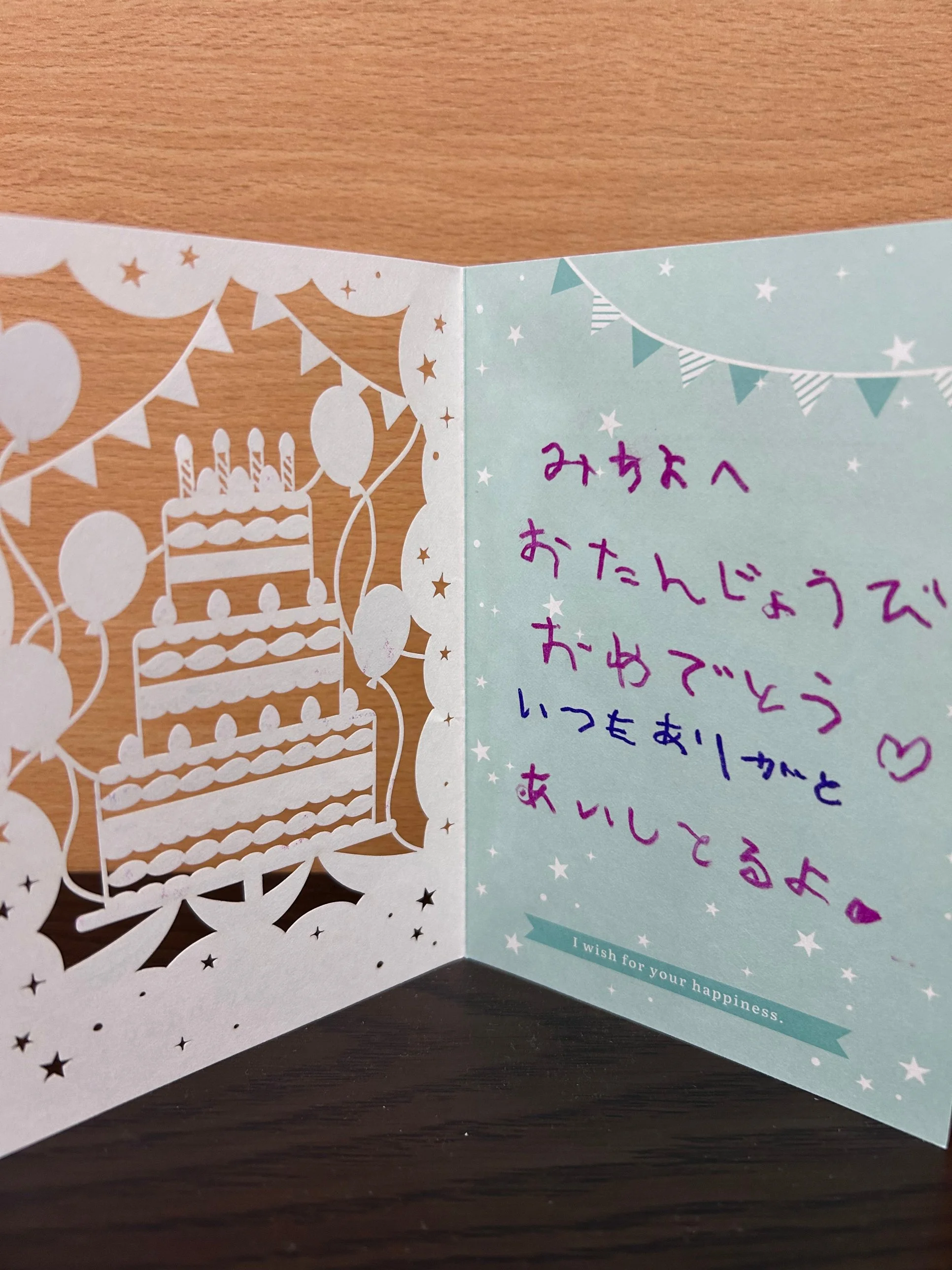 切り絵で3段のケーキや風船がデザインされたバースデーカード。右側のページに「おたんじょうび おめでとう いつもありがとう あいしてるよ」と手書きのメッセージが書かれている